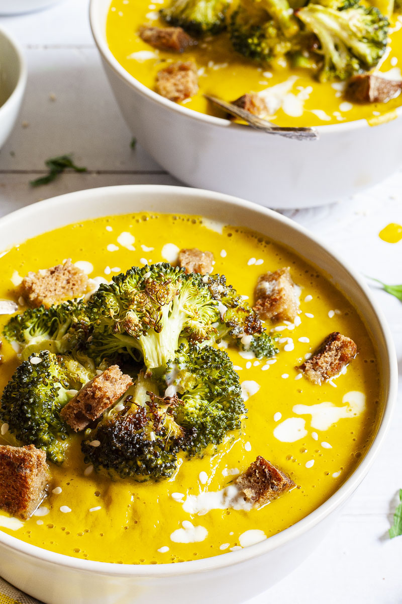 Yellow cream soup topped with roasted broccoli florets, brown croutons, white drizzle spots served in 2 white bowls with a spoon in each.  Other toppings like croutons, red pepper flakes and chopped parsley is in small bowls around the soups.