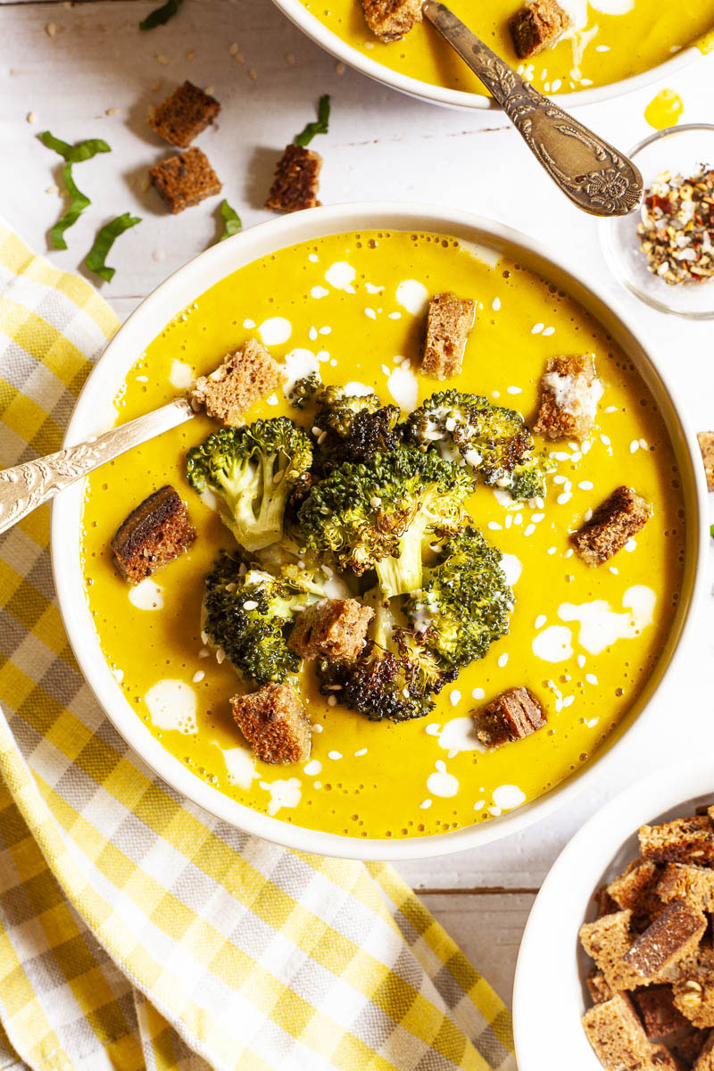 Yellow cream soup topped with roasted broccoli florets, brown croutons, white drizzle spots served in 2 white bowls with a spoon in each. Other toppings like croutons, red pepper flakes and chopped parsley is in small bowls around the soups.