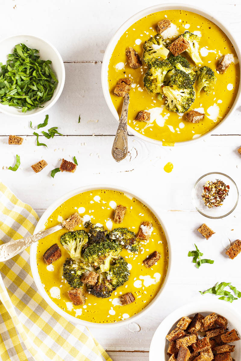 Yellow cream soup topped with roasted broccoli florets, brown croutons, white drizzle spots served in 2 white bowls with a spoon in each. Other toppings like croutons, red pepper flakes and chopped parsley is in small bowls around the soups.