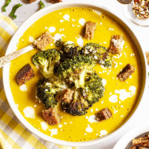 Yellow cream soup topped with roasted broccoli florets, brown croutons, white drizzle spots served in a white bowl with a spoon.