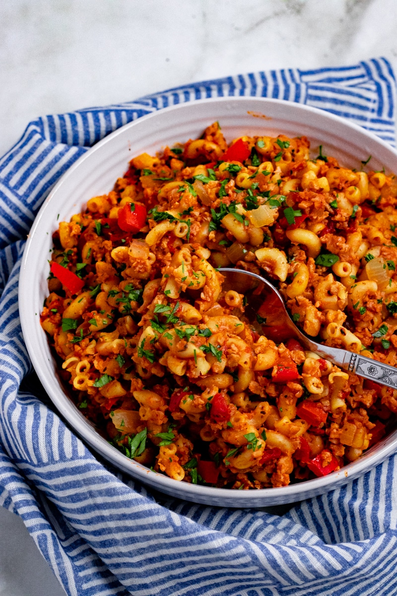 White bowl on blue tablecloth with elbow pasta, tomato chunks, fresh green herbs, and brown crumbles