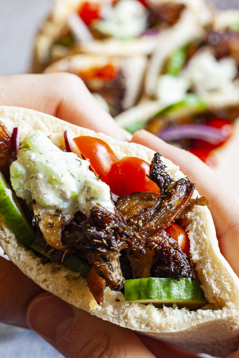 A pita pocket in a hand stuffed with crispy brown shredded oyster mushroom, tomato and cucumber slices and white-green tzatziki sauce