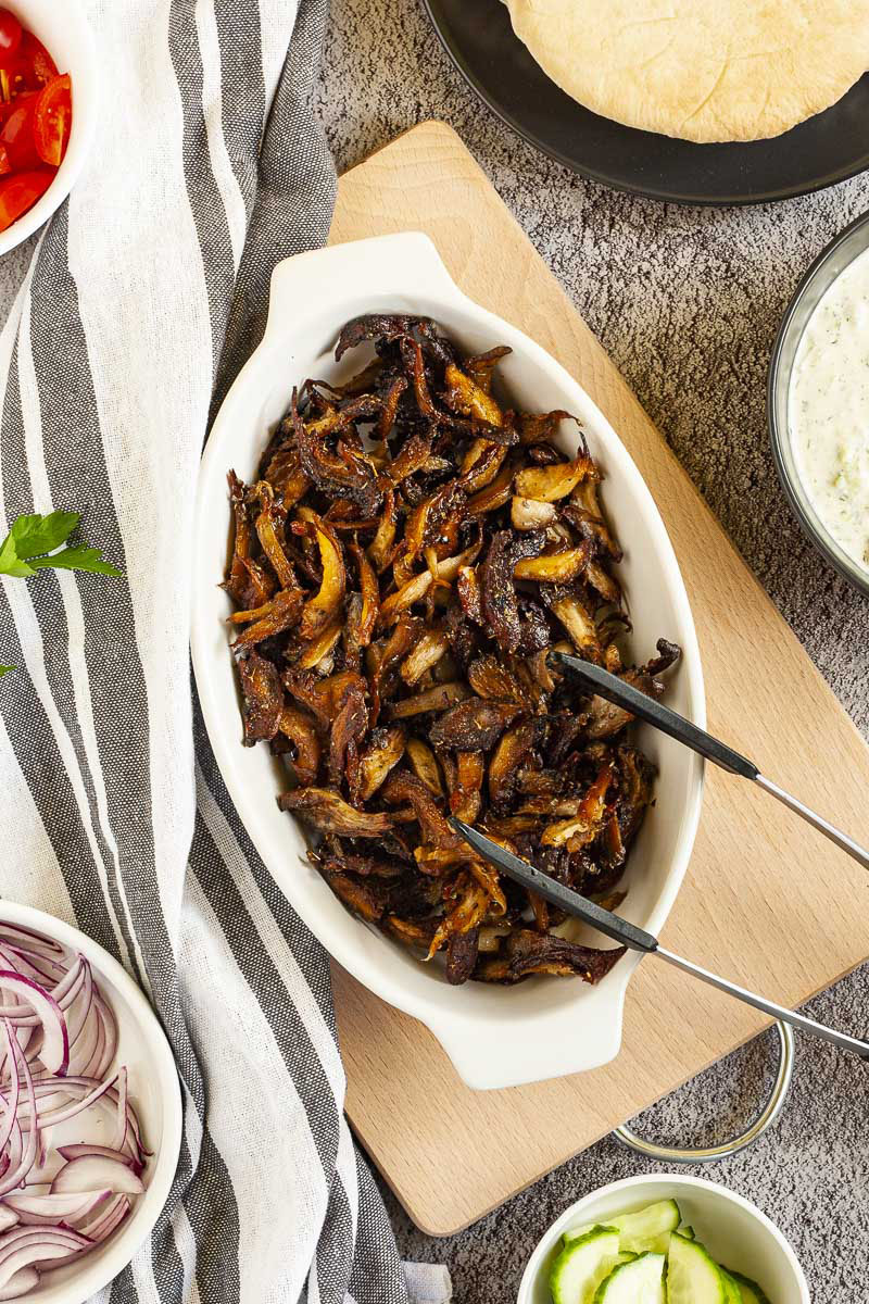 White serving bowl full of dark brown crispy shredded oyster mushrooms. Other ingredients are around in small bowls like purple onion, tomato and cucumber slices and pita bread.