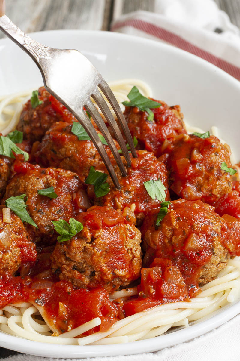 White serving bowl with several brown meatballs on top of spaghetti soaked in bright red tomato sauce. A fork is taking one ball from the middle.