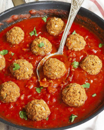 A skillet from above with several baked brown meatballs in bright red sauce and chopped green herbs. A spoon is taking one ball from the middle.