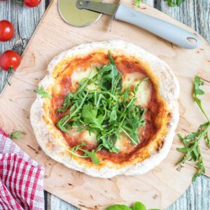 Pizza with fluffy crispy crust topped with tomato sauce, melted cheese and fresh arugula on a wooden board. Pizza cutter and more fresh herbs ingredients are around it.