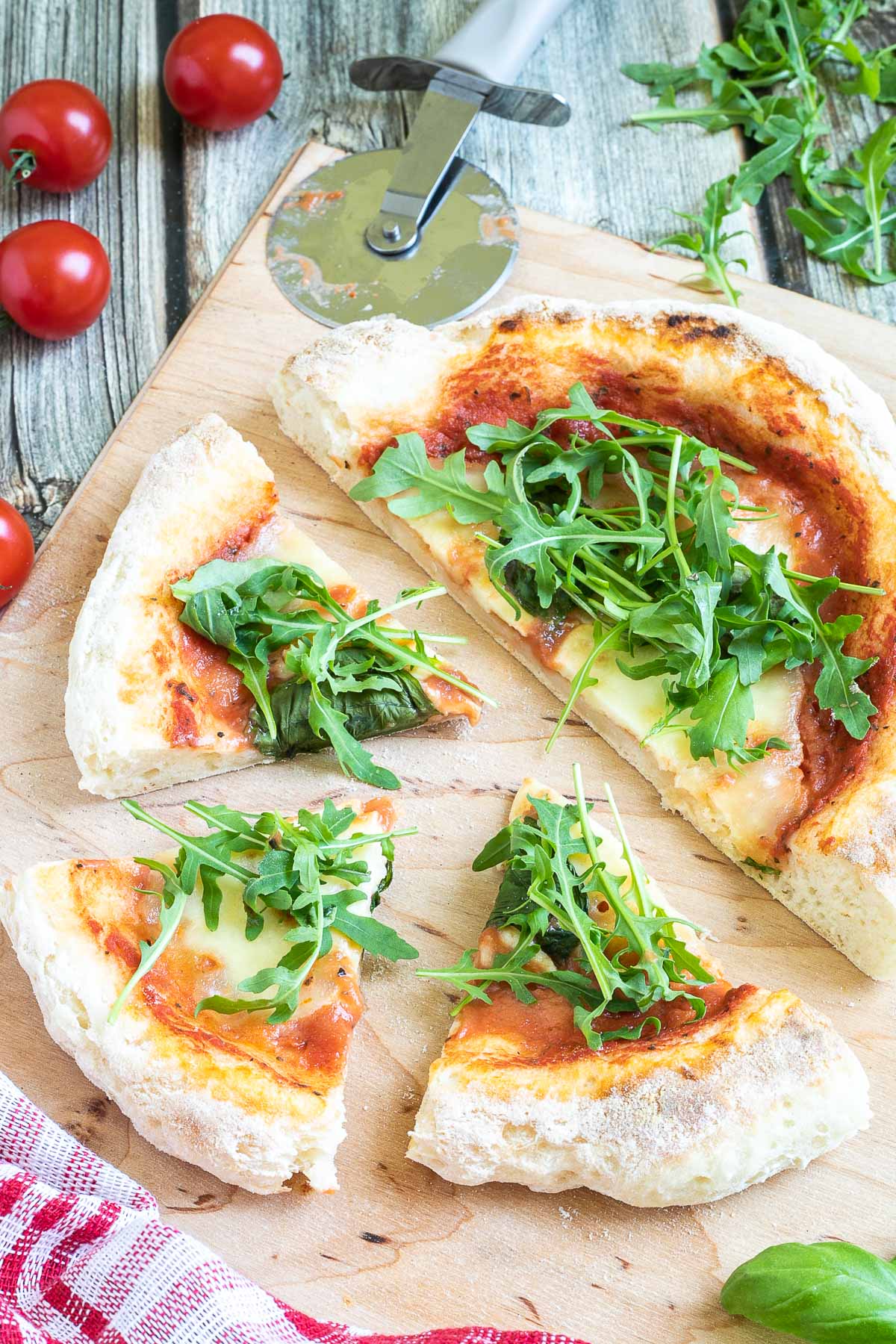 A pizza cut into slices with fluffy crispy crust topped with tomato sauce, melted cheese and fresh arugula on a wooden board. Pizza cutter and more fresh herbs ingredients are around it.