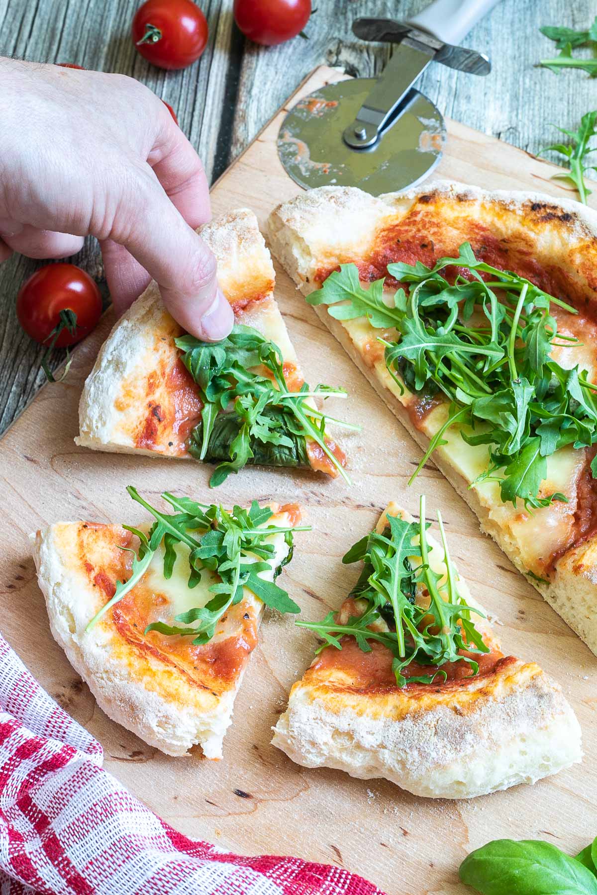 A pizza cut into slices with fluffy crispy crust topped with tomato sauce, melted cheese and fresh arugula on a wooden board. A hand is taking one slice.
