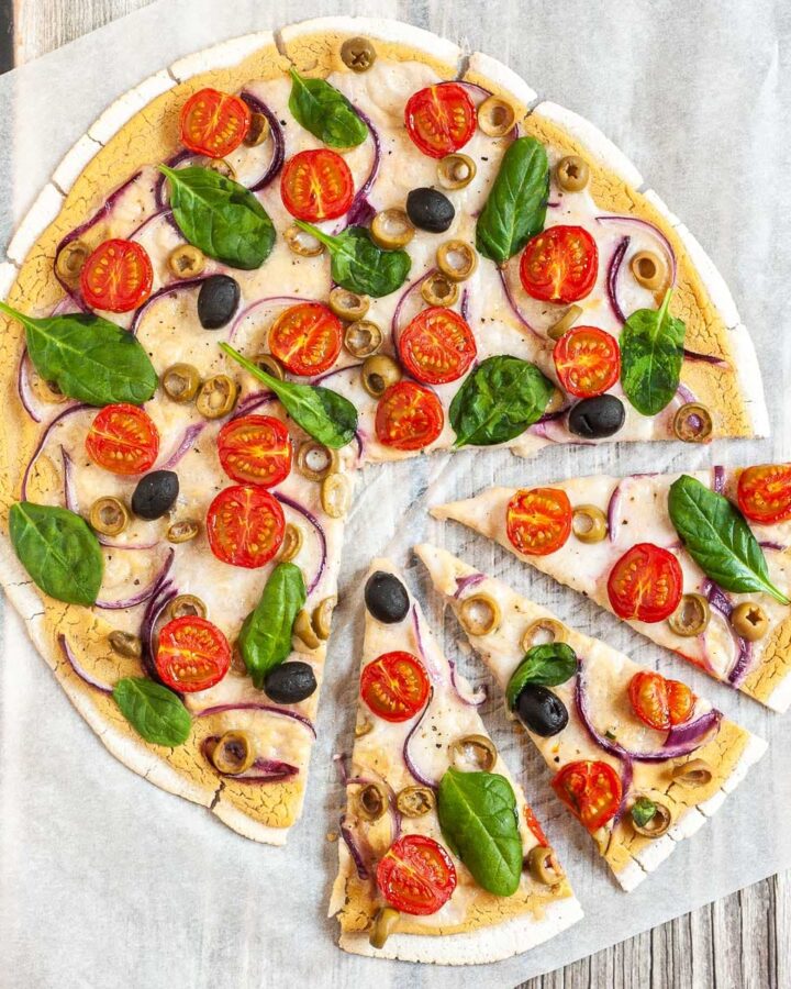Baked and sliced pizza from above on white parchment paper topped with hummus, cherry tomatoes, purple onion slices, green and black olives, melted cheese and fresh basil leaves.