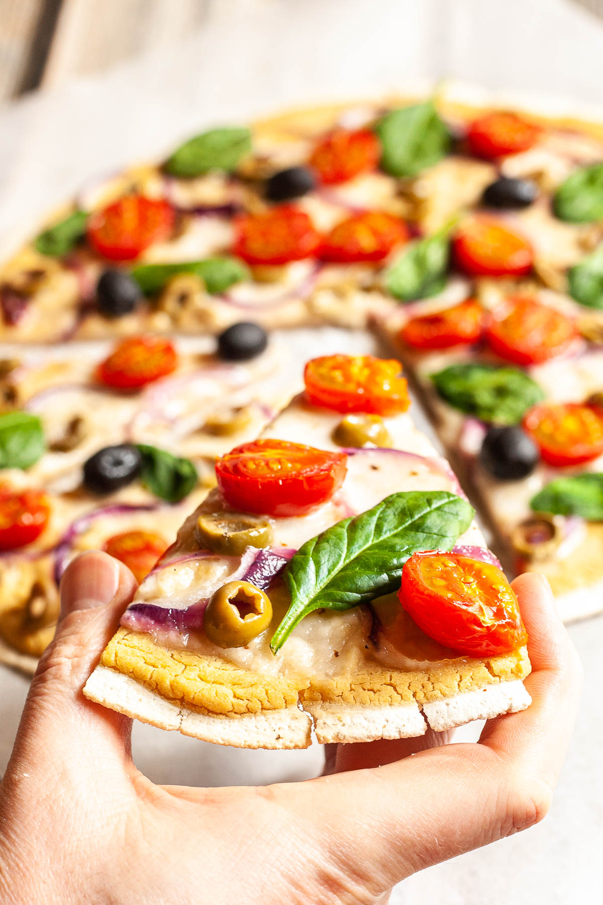 A hand is holding a slice of baked pizza with melted cheese, cherry tomatoes, purple onion slices, green olives and fresh basil leaves the remaining of the pizza is at the background.