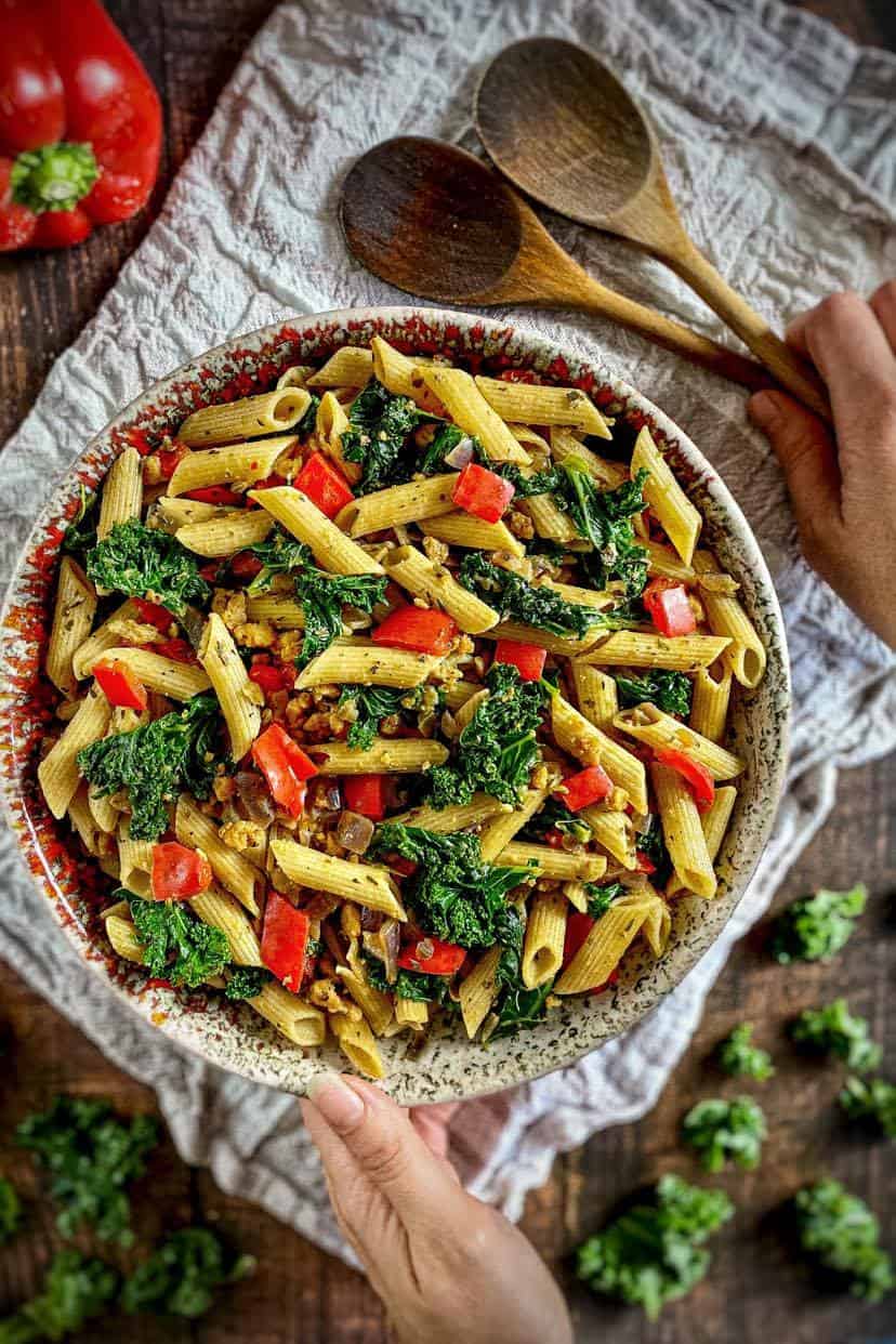 Large bowl with yellow penne pasta, kale pieces, red bell pepper slices and brown crumbles