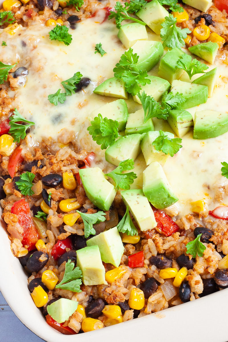 A ligth brown ceramic dish with colorful ingredients like black beans, corn, melted cheese, bell pepper strips, chopped avocado, chopped green herbs.