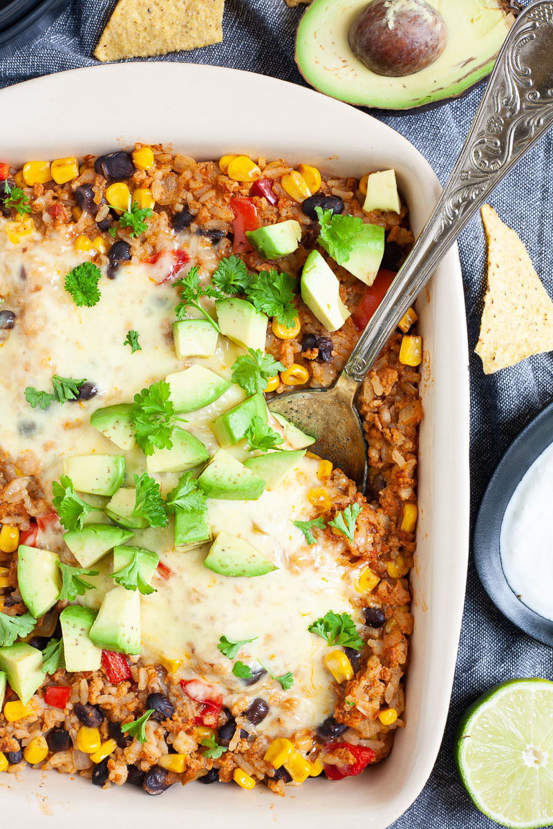 A ligth brown ceramic dish with colorful ingredients like black beans, corn, melted cheese, bell pepper strips, chopped avocado, chopped green herbs. A serving spoon is dunked in the middle. Half avocado, half lime and tortilla chips are scattered around