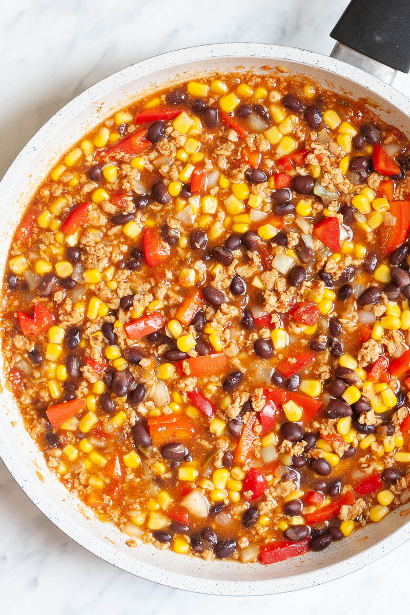 A white frying pan with colorful ingredients like black beans, corn, bell pepper strips, meat-like crumbles in a red sauce.