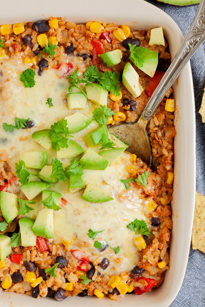 A ligth brown ceramic dish with colorful ingredients like black beans, corn, melted cheese, bell pepper strips, chopped avocado, chopped green herbs. A serving spoon is dunked in the middle.