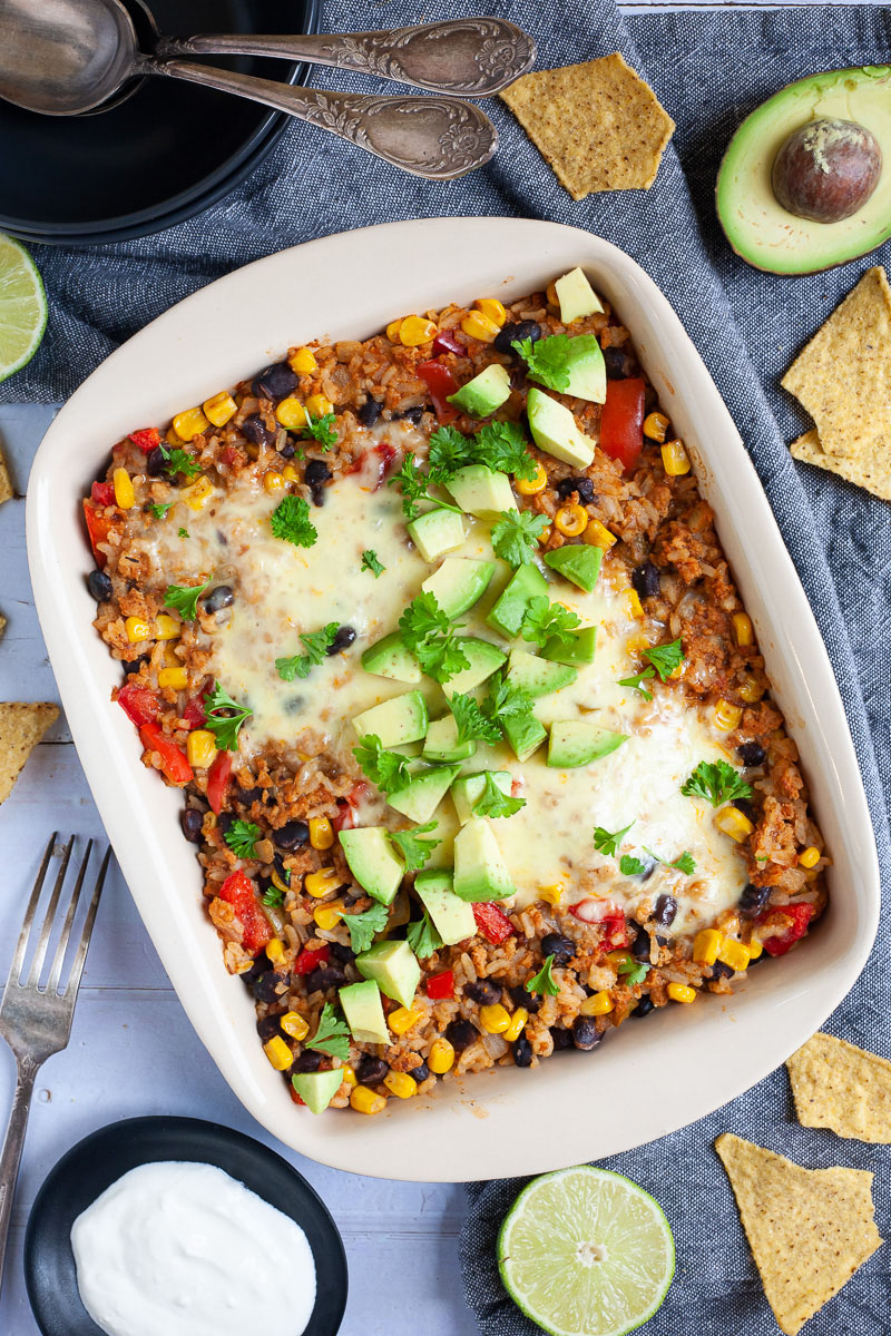 A ligth brown ceramic dish with colorful ingredients like black beans, corn, melted cheese, bell pepper strips, chopped avocado, chopped green herbs. A serving spoon is dunked in the middle. Half avocado, half lime and tortilla chips are scattered around.