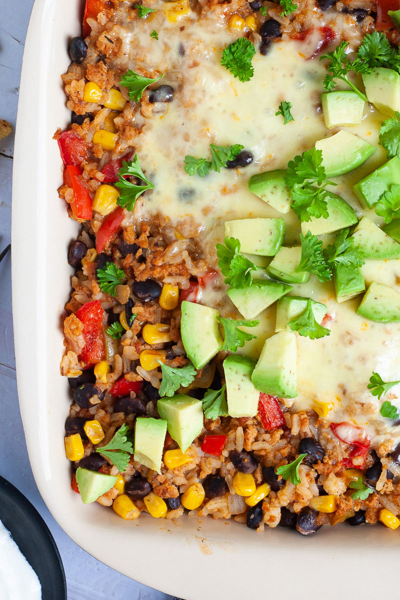 A ligth brown ceramic dish with colorful ingredients like black beans, corn, rice, melted cheese, bell pepper strips, chopped avocado, chopped green herbs.