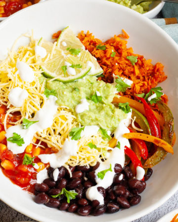 A large white plate in the middle with colorful ingredients like black beans, corn, shredded cheese, bell pepper strips, lime, orange rice. Small white bowl are scattered around with the same ingredients.