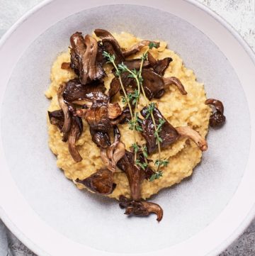 White bowl from above with creamy yellow polenta topped with brown mushroom shreds and fresh herbs