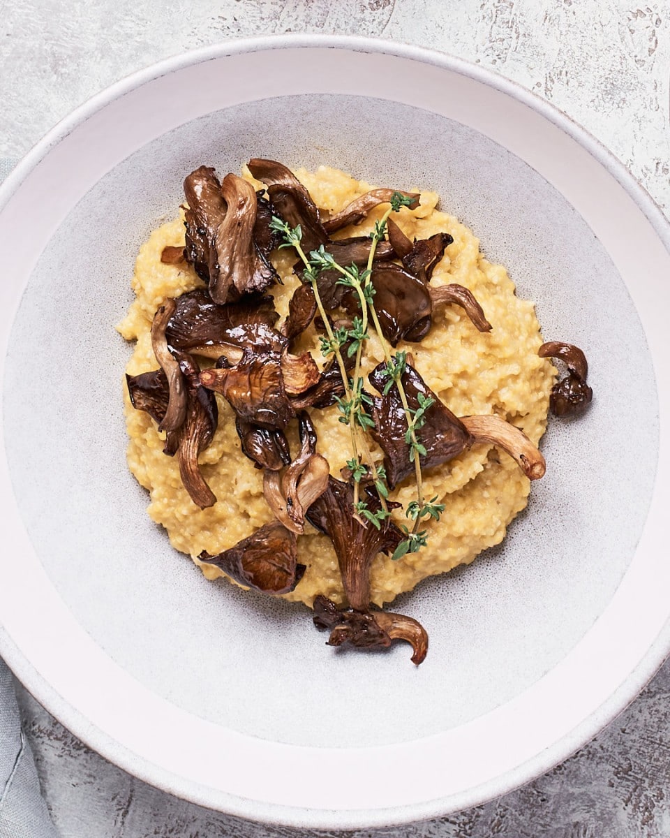 White bowl from above with creamy yellow polenta topped with brown mushroom shreds and fresh herbs