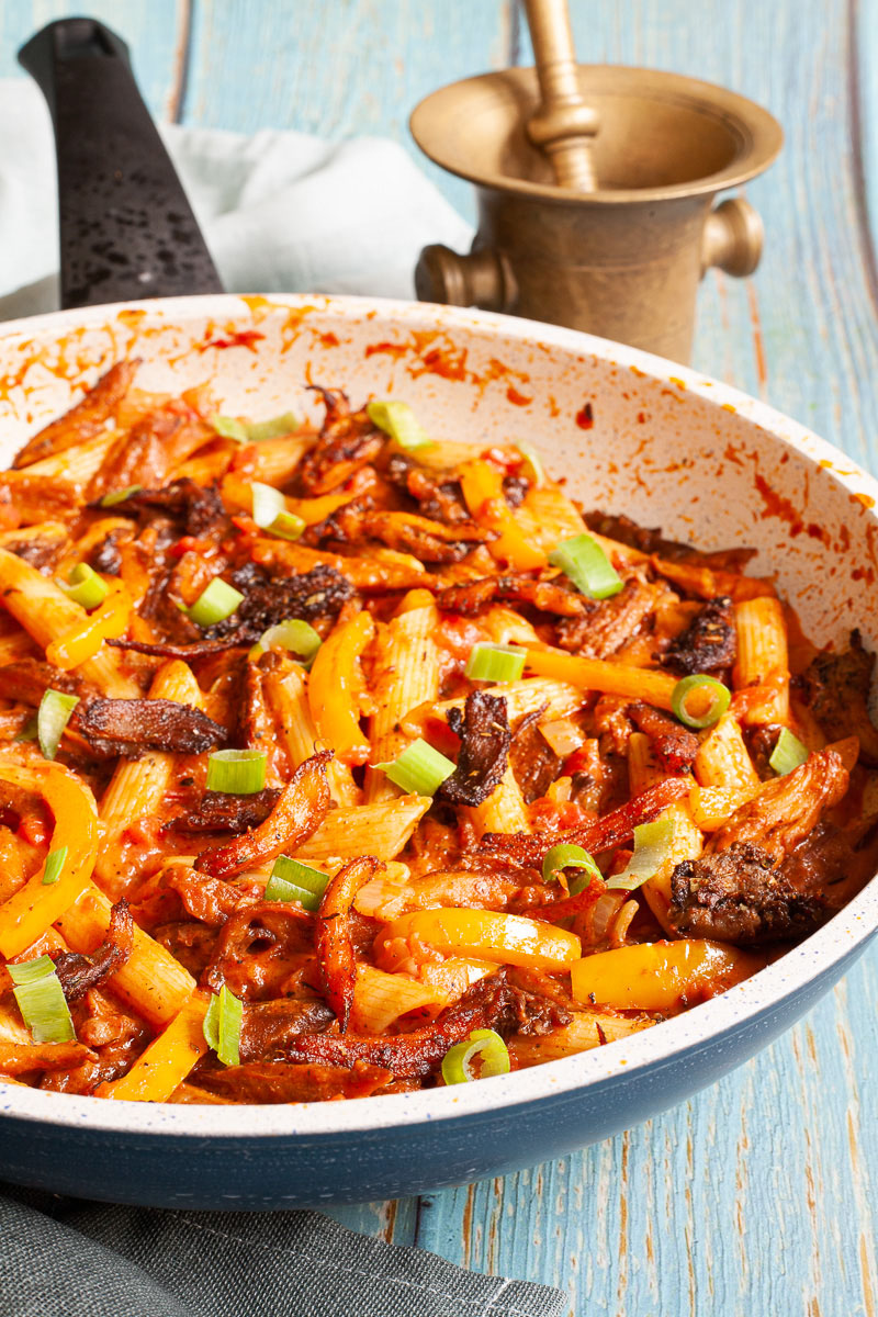 Penne pasta with yellow bell pepper slices, brown mushroom shreds and spring onion rings in a creamy red sauce in a large frying pan