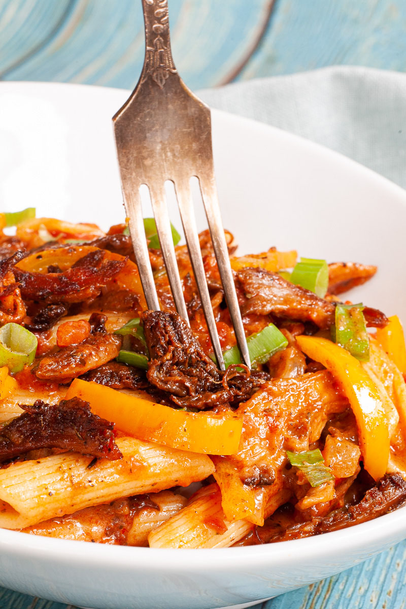 Penne pasta with yellow bell pepper slices, brown mushroom shreds and spring onion rings in a creamy red sauce served in a white bowl. A fork is piercing through the middle.