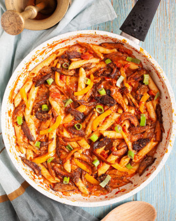 Penne pasta with yellow bell pepper slices, brown mushroom shreds and spring onion rings in a creamy red sauce in a frying pan from above.
