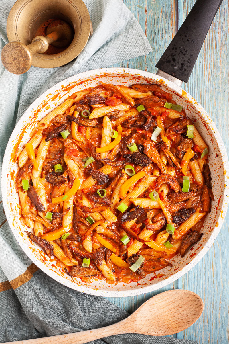 Penne pasta with yellow bell pepper slices, brown mushroom shreds and spring onion rings in a creamy red sauce in a frying pan from above.