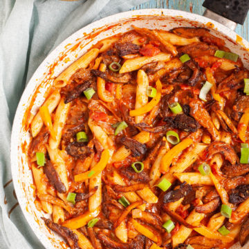 Penne pasta with yellow bell pepper slices, brown mushroom shreds and spring onion rings in a creamy red sauce in a frying pan from above.