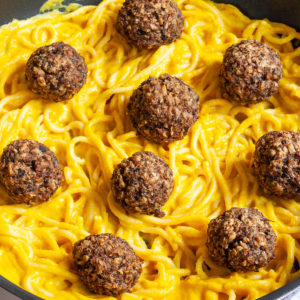 Several mushroom meatballs on top of spaghetti with yellow creamy sauce served on a black plate. A fork is taking on meatballs from the middle.