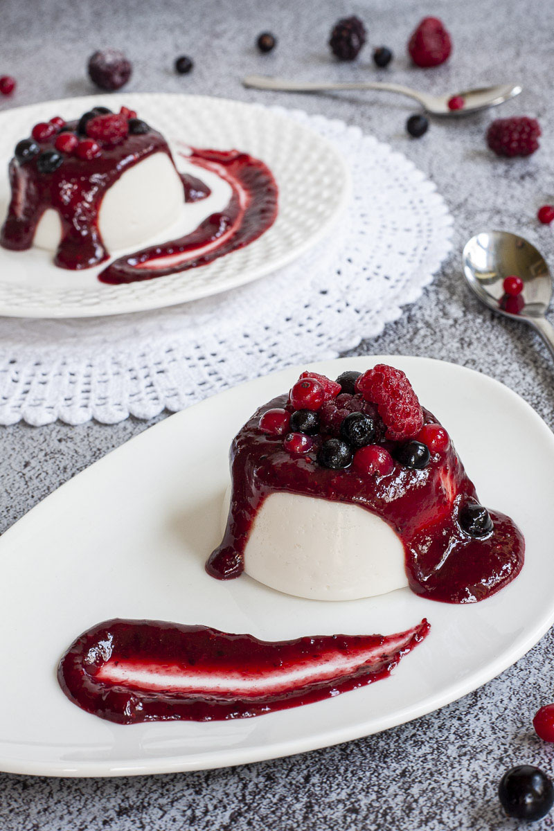 2 white plates with white panna cotta topped with a thick purple sauce and different berries.