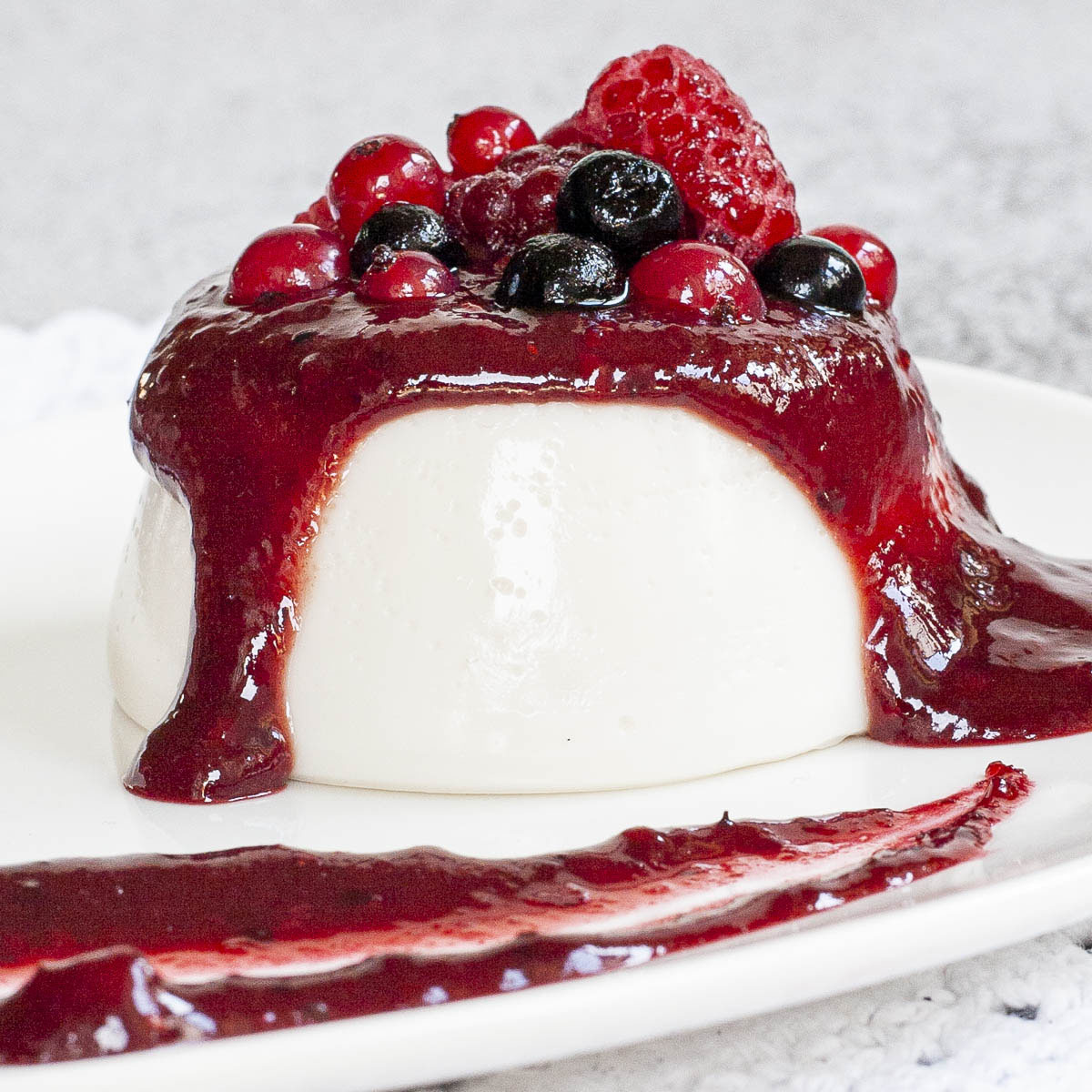 A white plate with white panna cotta topped with a thick purple sauce and different berries up really close.