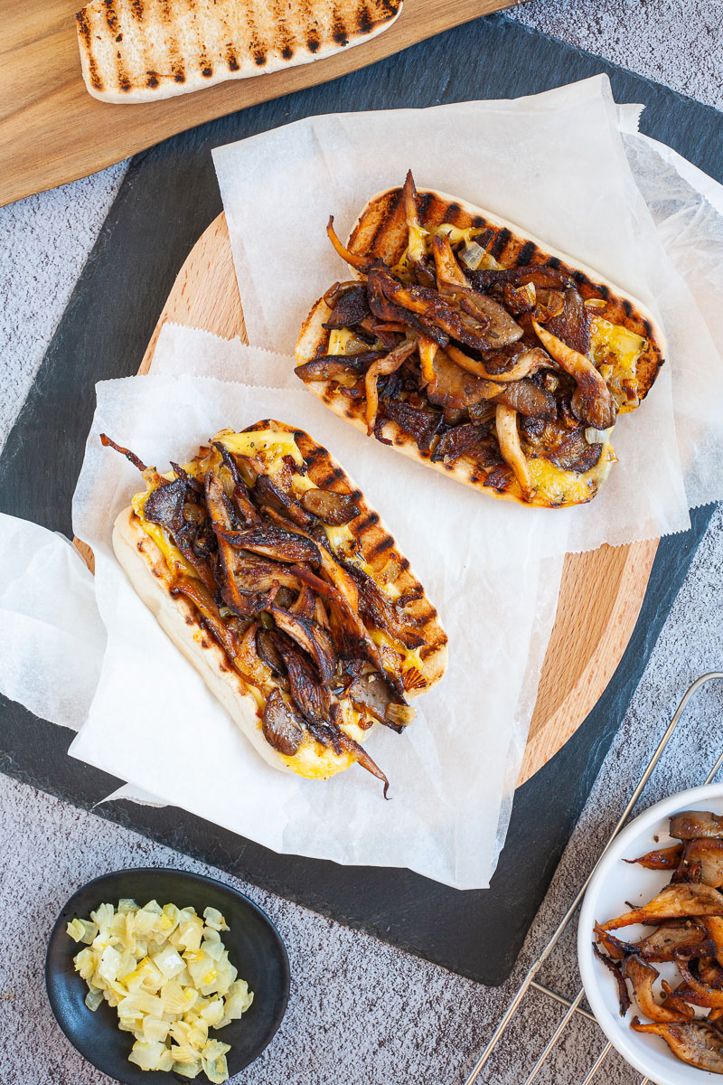 2 bread loaves stuffed with brown mushroom shreds and melted cheese wrapped in a white paper. A small white bowl with extra mushroom shreds and a small black bowl with extra yellow onion pieces are next to them.