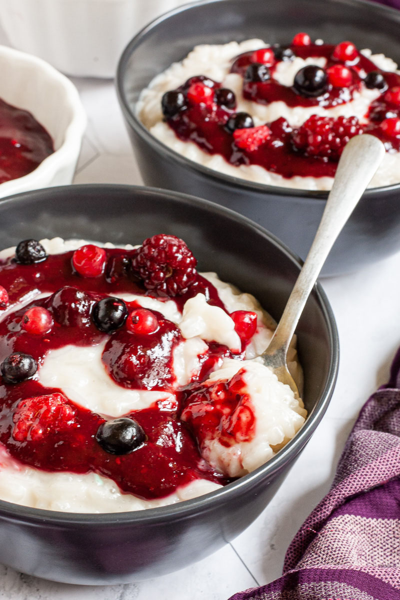 A black bowl up close with white pudding topped with a swirl of thick red-purple sauce and different berries. A spoon is taking the first bite. Another bowl is in the background.