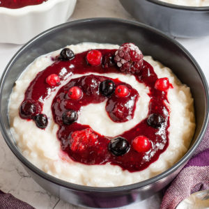 A black bowl up close with white rice pudding topped with a swirl of thick red-purple sauce and different berries.