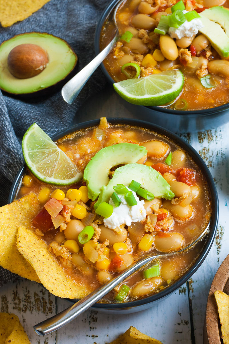 2 black bowls with orange liquid, beans, corn, chopped tomaotes, mince, avocado slices, chopped spring onion, lime slices, and tortilla chips. Half of avocado and more tortilla chips are scattered around