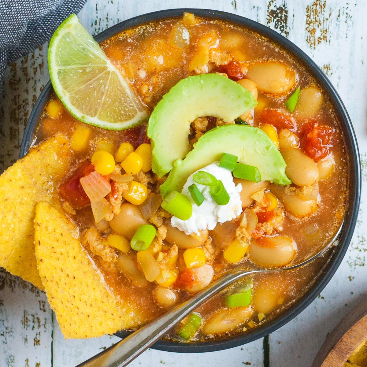 A black bowl with orange liquid, beans, corn, chopped tomaotes, mince, avocado slices, chopped spring onion, lime slices, and tortilla chips.