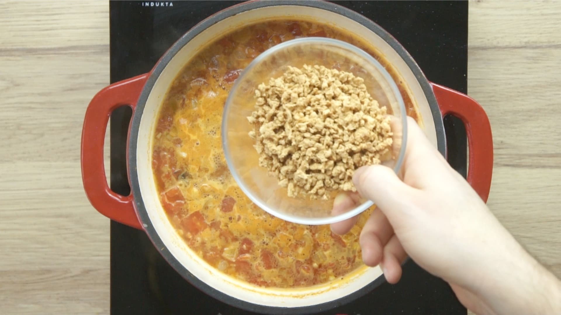 A hand is holding a bowl of crumbles to be added to a tomato colored soup below.