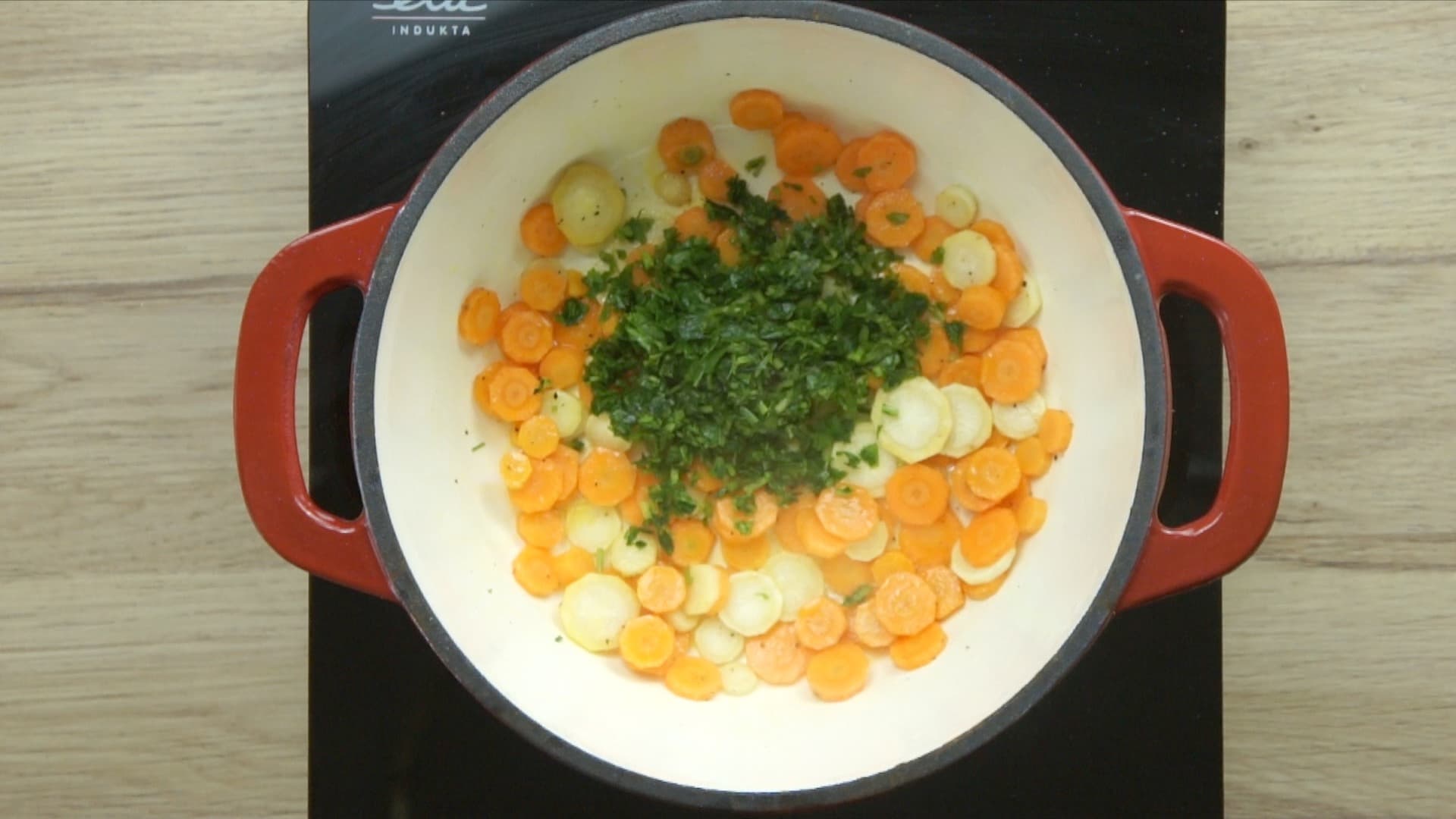 Red enameled Dutch oven with carrot slices, parsnip slices, and freshly chopped green herbs.