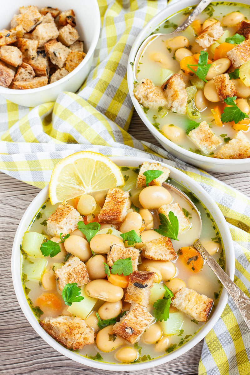 Light yellow soup served in two white bowls full of large white beans, croutons, carrot slices, and diced potatoes. It is sprinkled with freshly chopped herbs.