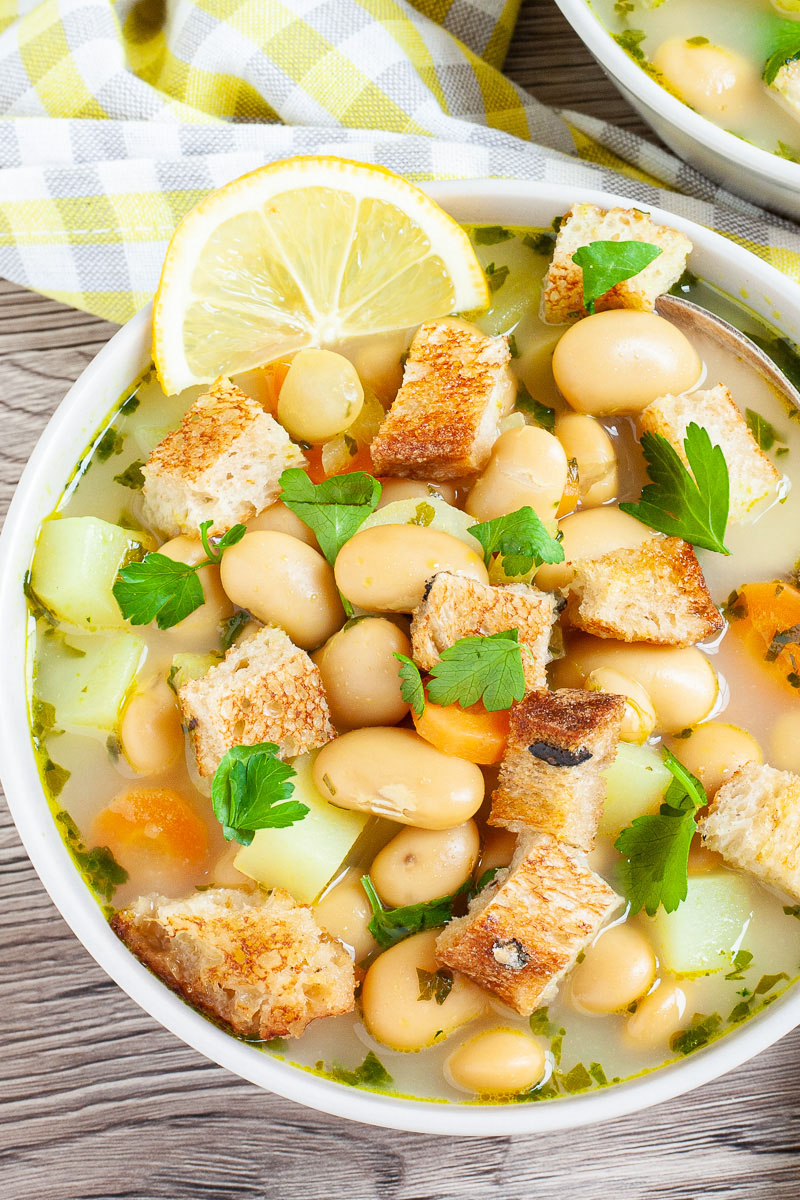 Light yellow soup served in a white bowl with a spoon full of large white beans, croutons, carrot slices, diced potatoes and freshly chopped herbs.