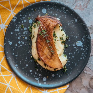 A slice of dark brown grilled king oyster mushrooms on top of a yellow mash served on a black plate.