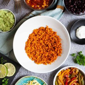 A large white plate in the middle orange riced sweet potato. Small white bowl are scattered around with colorful ingredients like black beans, corn, shredded cheese, bell pepper strips, lime, orange rice.