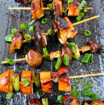 Wooden skewers with mushroom slices grilled to dark brown almost black on the edges. They are sprinkled with green onion rings