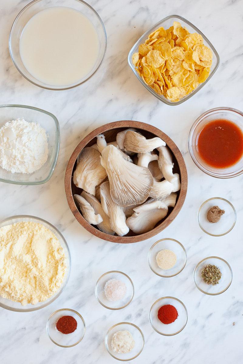 The ingredients to make vegan chicken wings are placed in several glass bowls: milk, flour, corn flakes, oyster mushrooms, hot sauce, different spices and herbs