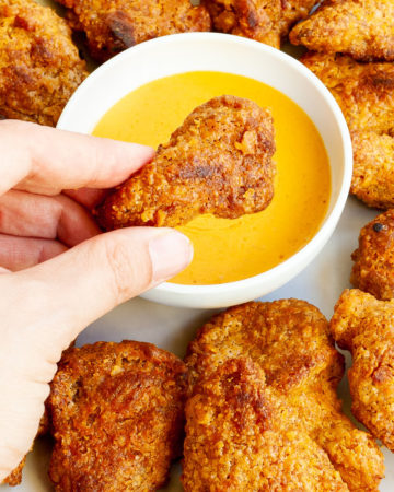Brown breaded wings on a white plate with a yellow sauce in the middle. A hand is holding one piece