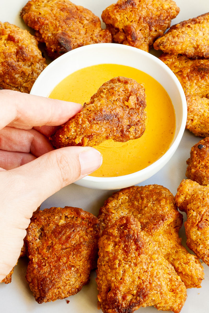 Brown breaded wings on a white plate with a yellow sauce in the middle. A hand is holding one piece