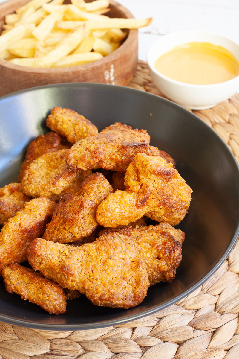 Brown breaded wings in a black bowl. French fries next to it in a wooden bowl and a yellow sauce in a small white bowl.