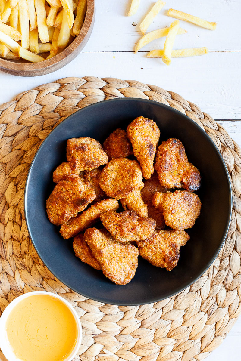 Brown breaded wings in a black bowl. French fries next to it in a wooden bowl and a yellow sauce in a small white bowl.