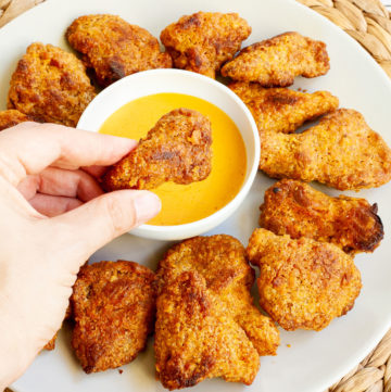 Brown breaded wings on a white plate with a yellow sauce in the middle. A hand is holding one piece
